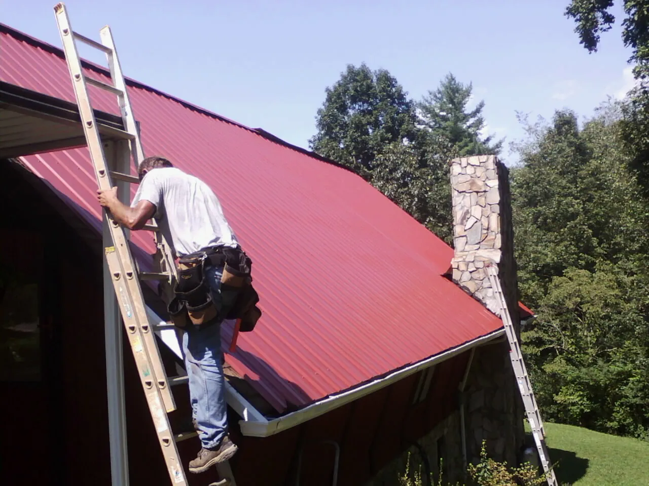 Roofer on a ladder beside a red metal roof and stone chimney — Soffit & Fascia Repair by Roofing Brothers in Staffordsville